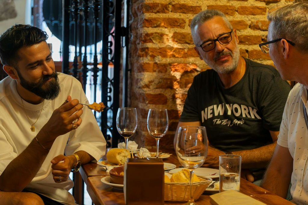 빵, 와인잔, 접시를 들고 테이블에 앉아 벽돌 벽 앞에서 대화를 나누는 두 명의 남성.