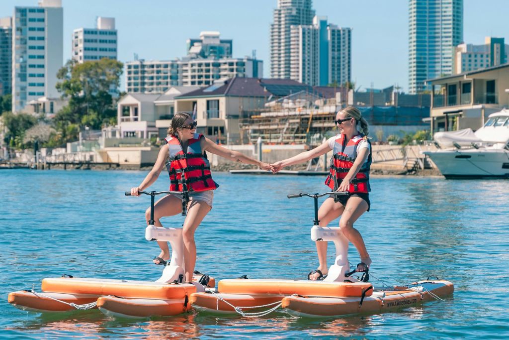 구명조끼를 입은 두 사람이 차분한 물가에서 수상 자전거를 타고 있으며, 도시 건물이 배경으로 보입니다.