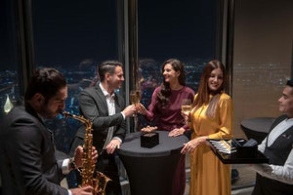 고층 바에서 음료를 즐기는 우아한 옷차림의 네 명. 웨이터가 시가 트레이를 제공합니다. 창문으로 보이는 도시의 불빛.