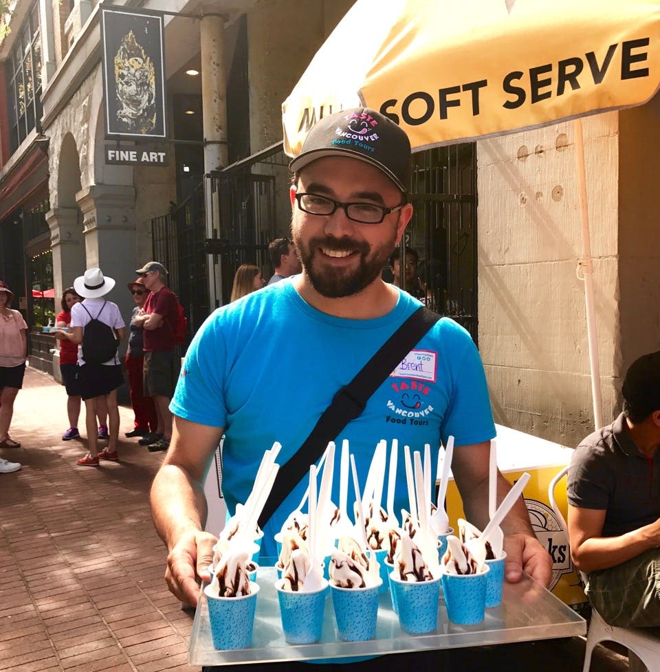 한 남성이 초콜릿 소스와 화이트 스푼을 얹은 소프트 아이스크림이 담긴 파란색 컵 쟁반을 들고 미소를 짓고 있습니다.