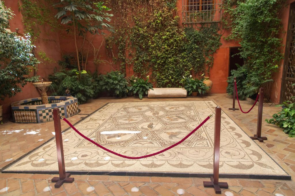 식물과 밧줄로 둘러싸인 화려한 모자이크 바닥으로, 주황색 벽과 녹지가 있는 안뜰에 자리잡고 있습니다.