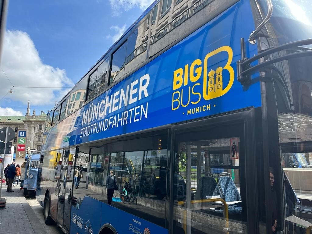 '뮌헨 슈타트룬트파르텐 (Münchener Stadtrundfahrten)' 과 '빅 버스 뮌헨 (Big Bus Munich)' 이라는 이름의 파란색 2층 버스는 도심 거리 근처에 주차되어 있으며, 내부에 사람들이 보입니다.