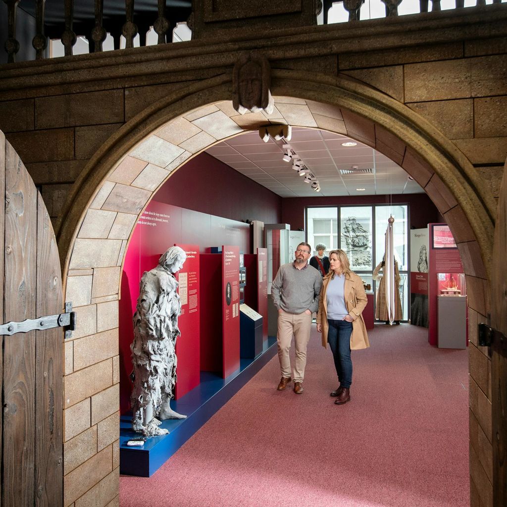 나무 문이 있는 석조 아치형 통로를 통해 바라보는 동상과 정보 디스플레이가 특징인 박물관 전시관의 두 사람.