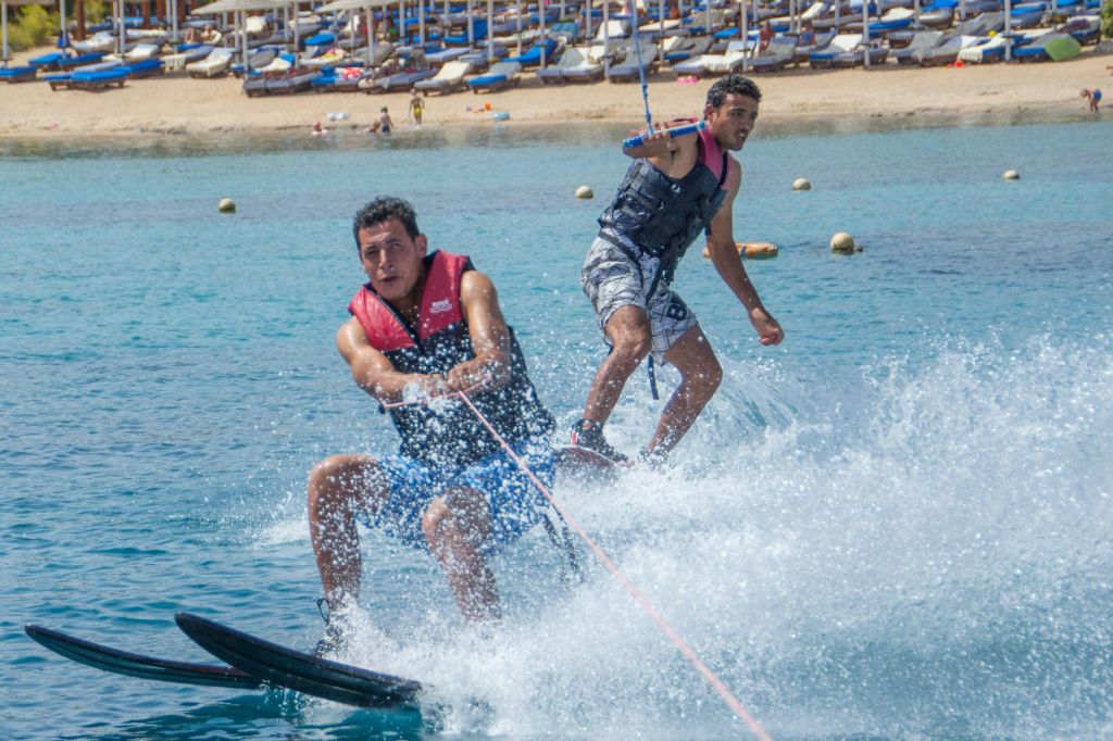 해안가에서 구명조끼를 입은 두 명의 남성 수상스키. 일광욕 의자와 사람들이 있는 모래 해변이 배경으로 보입니다.