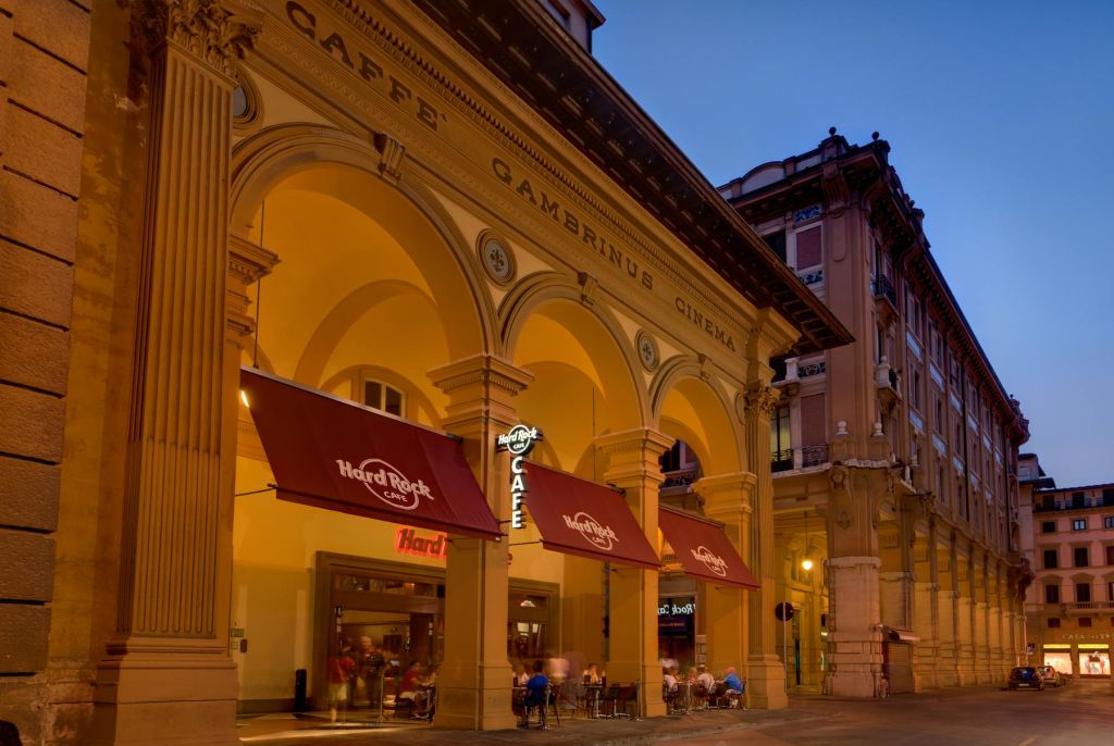 아치와 "Caffè Gambrinus Cinema" 간판이 있는 조명 카페입니다. 빨간색 "Hard Rock Café" 차양 아래에 사람들이 앉아 있습니다.