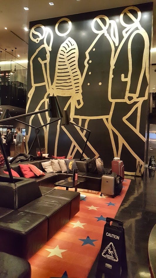 검은 가죽 의자, 베개, 여행 가방, 추상적인 인물이 그려진 커다란 벽 벽화가 있는 세련된 라운지 공간입니다.