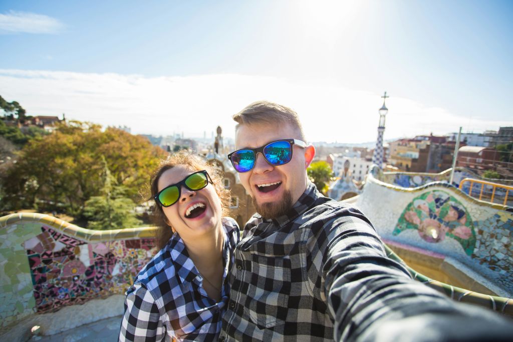 선글라스를 쓰고 체크무늬 셔츠를 입은 미소 짓는 남녀가 햇살 가득한 공원에서 도시 풍경과 모자이크 예술을 배경으로 셀카를 찍고 있습니다.