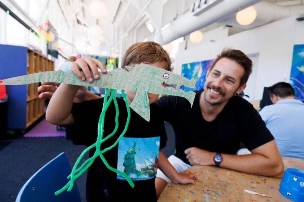 한 어린이가 미술실에서 웃는 어른 옆에 앉아 악어를 닮은 수제 종이 공예품을 들고 있습니다.