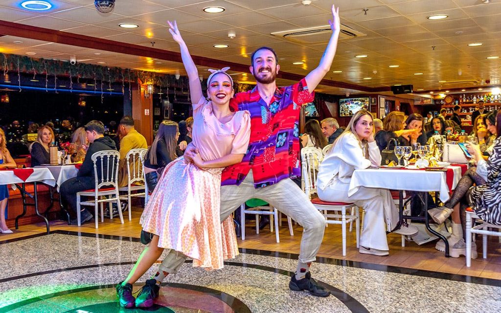 두 명의 공연자가 레스토랑에서 춤을 추고 있으며, 좌석에 앉아 후원자들이 지켜보고 있습니다. 숙소는 활기찬 분위기로 조명이 밝습니다.