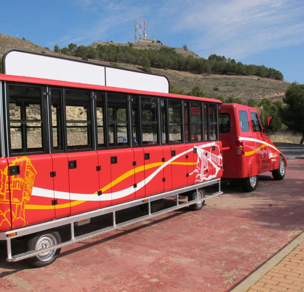 파란 하늘 아래 언덕과 나무를 배경으로 포장된 공간에 검은색 창문이 있는 빨간색 관광 트램.