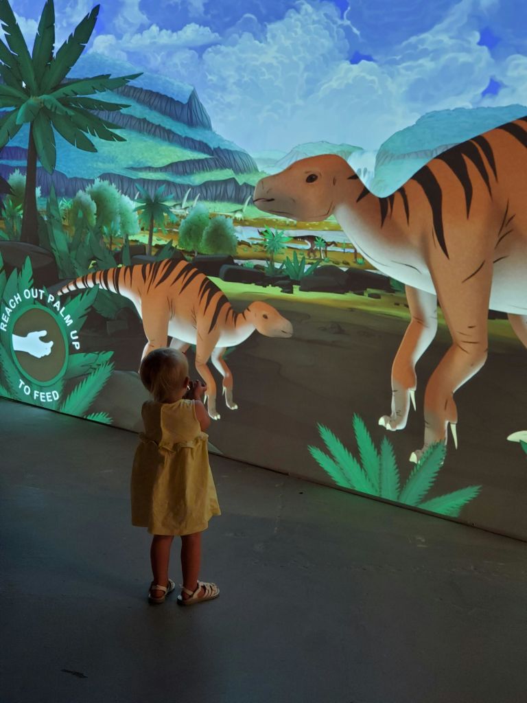 노란색 드레스를 입은 어린이가 울창한 선사 시대 풍경 속에서 공룡의 디지털 디스플레이와 상호 작용합니다. 표시된 텍스트에는 "손바닥을 뻗어 먹이를 주세요." 라고 쓰여 있습니다.