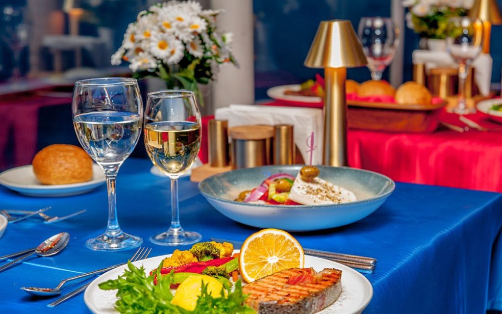 와인잔, 연어구이, 야채, 샐러드가 놓인 테이블입니다. 배경에는 꽃과 작은 램프가 있습니다.