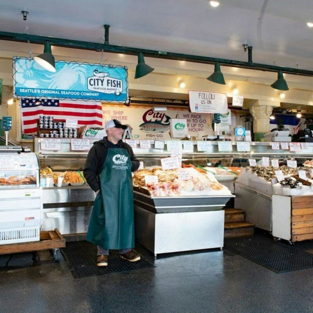 녹색 앞치마를 두른 사람이 해산물 카운터 앞에 서서 시장에 다양한 물품을 전시하고 있습니다.