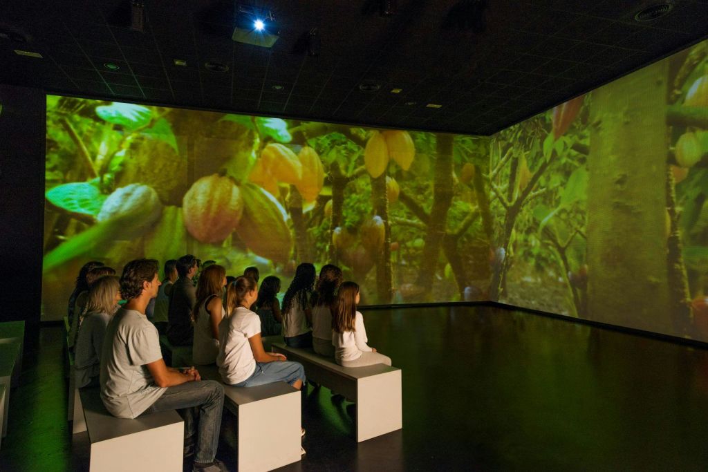 사람들은 어두운 방의 벤치에 앉아 나무에 커다란 열매가 그려진 커다란 몰입형 비디오를 보았습니다.