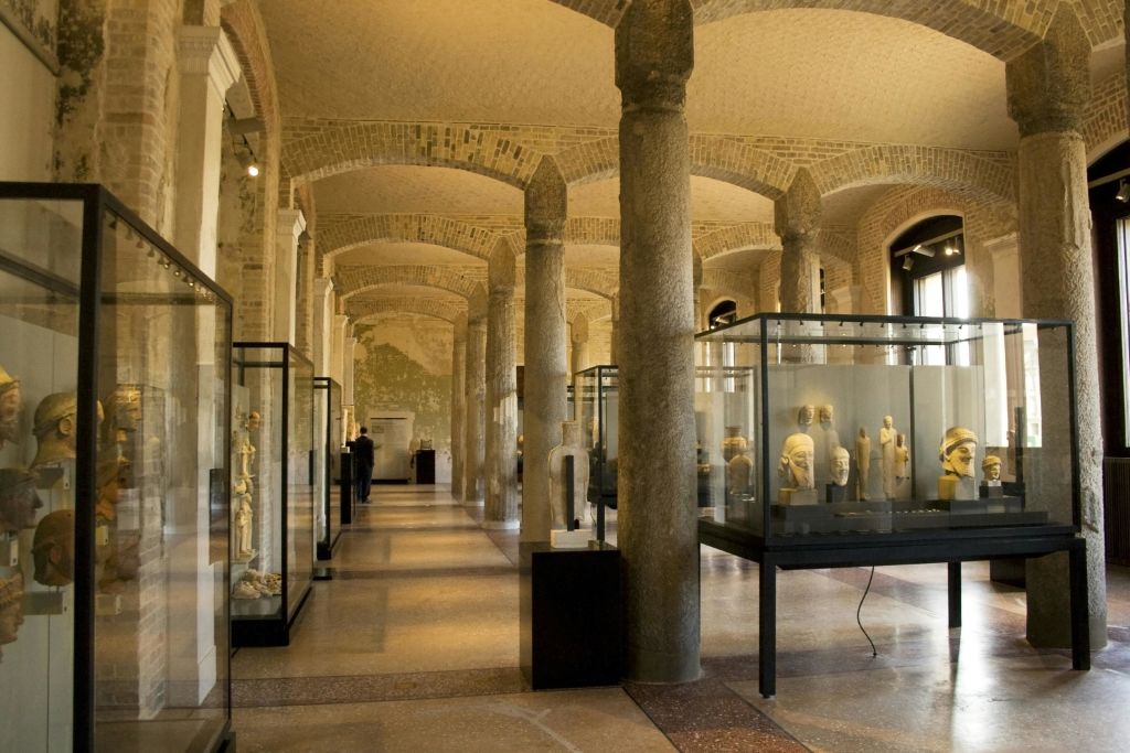 석조 기둥, 아치형 천장, 유리 진열 케이스로 조각과 유물을 전시하는 박물관 홀입니다.