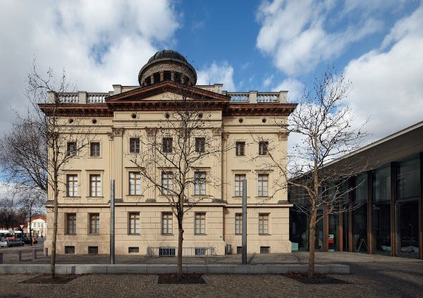 돔, 커다란 창문, 맨발의 나무, 부분적으로 흐린 하늘 아래 자갈길이 있는 고전적인 베이지색 건물입니다.