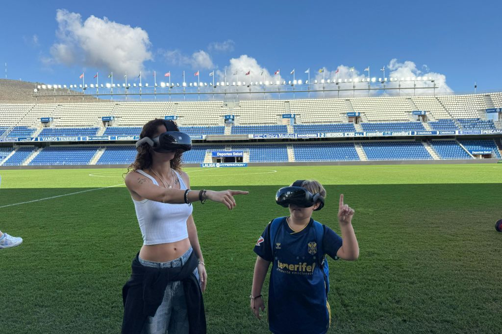 VR 헤드셋을 착용한 두 사람이 축구장에 서 있습니다. 경기장 좌석과 밝은 하늘이 배경에 있습니다.