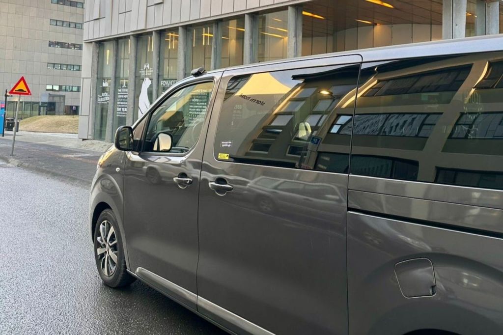 커다란 창문과 내부 조명이 있는 현대적인 건물 앞 거리에 회색 미니밴이 주차되어 있습니다.