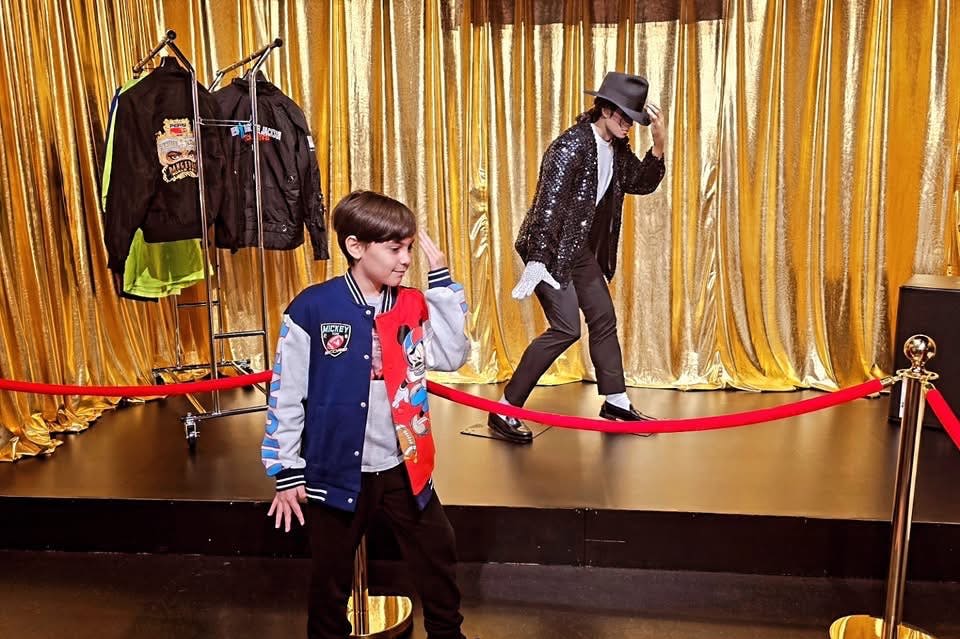 한 소년이 시퀸 재킷과 모자를 입은 마네킹 앞에서 댄스 포즈를 취하고 뒤에는 재킷이 전시되어 있습니다.