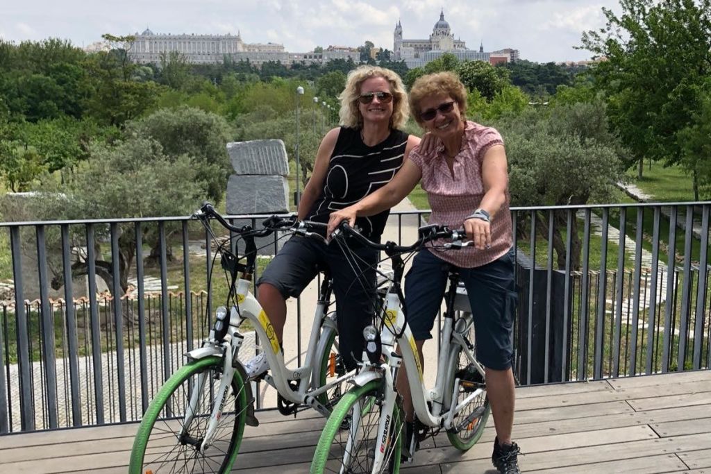 아름다운 공원과 유서 깊은 건물을 배경으로 다리 위에서 자전거를 탄 두 여성이 미소를 짓고 있습니다.