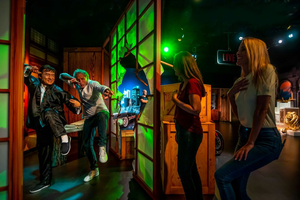 테마가 있는 전시관에서 부서진 벽을 닮은 세트 앞에서 두 사람이 무술 포즈를 취하고 있다. 한 여성이 근처를 지켜보고 있다.