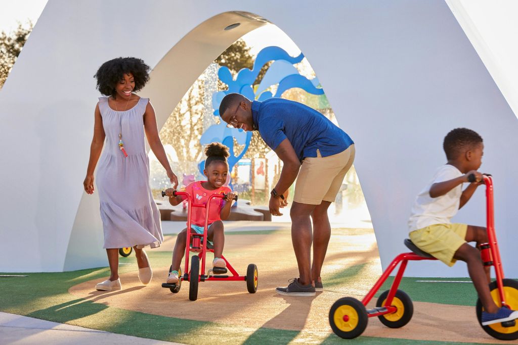 한 어린 소녀가 세발자전거를 타는 동안 한 여성과 한 남성이 장난기 가득한 야외 공간의 아치 아래에서 미소를 지으며 그녀를 관찰합니다.