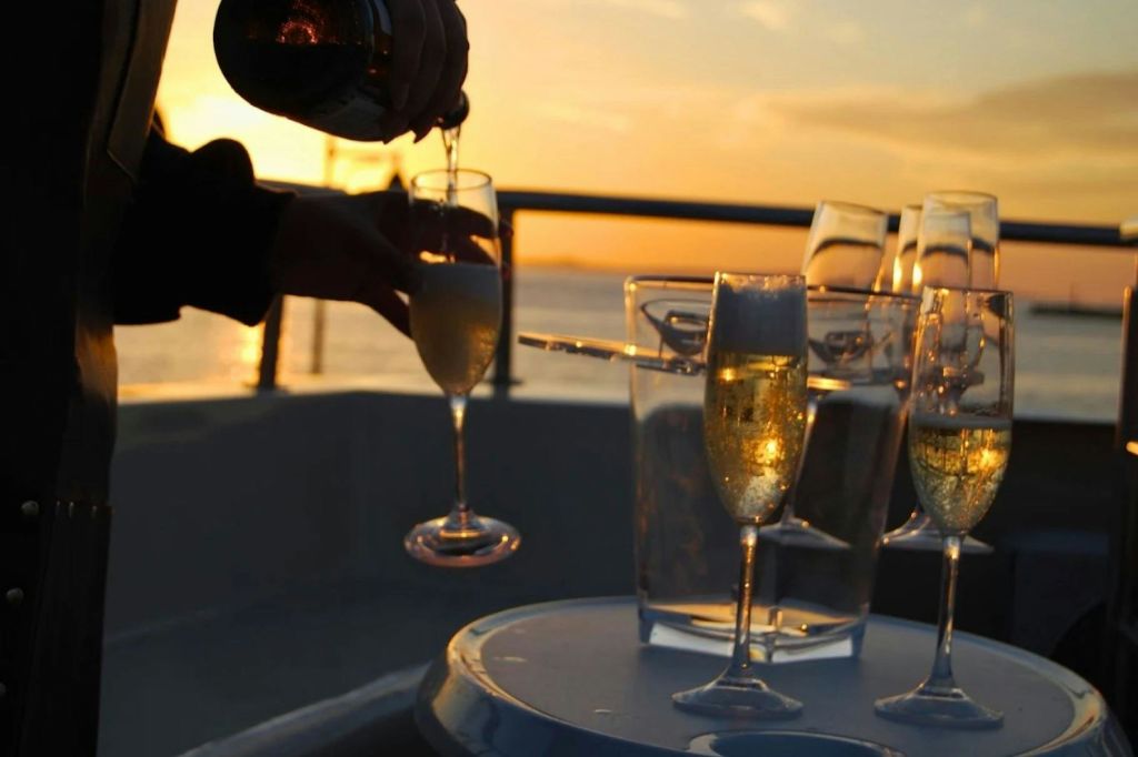 더 많은 유리잔과 얼음 양동이가 놓인 테이블 위의 유리잔에 샴페인을 담아 물 위로 지는 석양을 배경으로 손으로 샴페인을 부어줍니다.