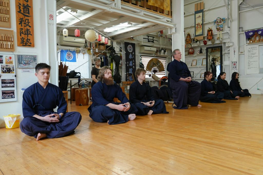 무술 유니폼을 입은 세 명의 남성이 도장에서 나무 바닥에 다리를 꼬고 앉아 명상을 하고 있습니다.