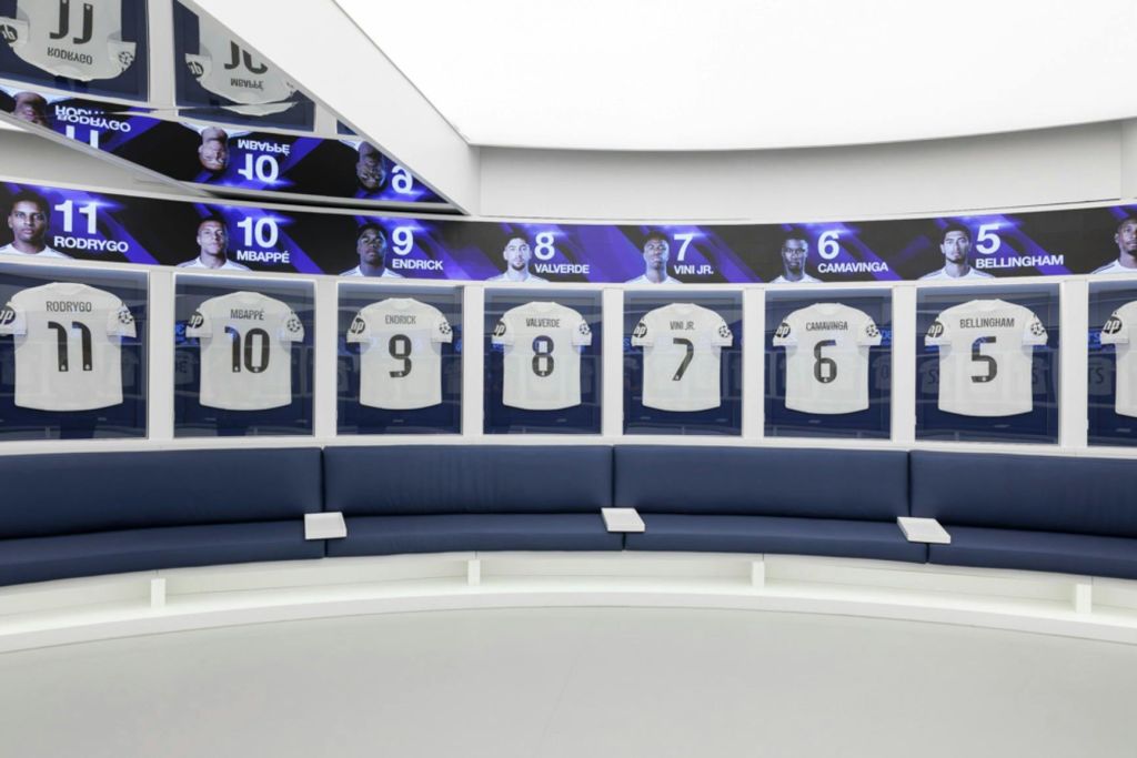 스포츠 선수의 번호가 매겨진 유니폼을 보여주는 디스플레이 케이스가 있는 현대적인 라커룸입니다. 파란색 좌석은 반원형으로 배열되어 있습니다.