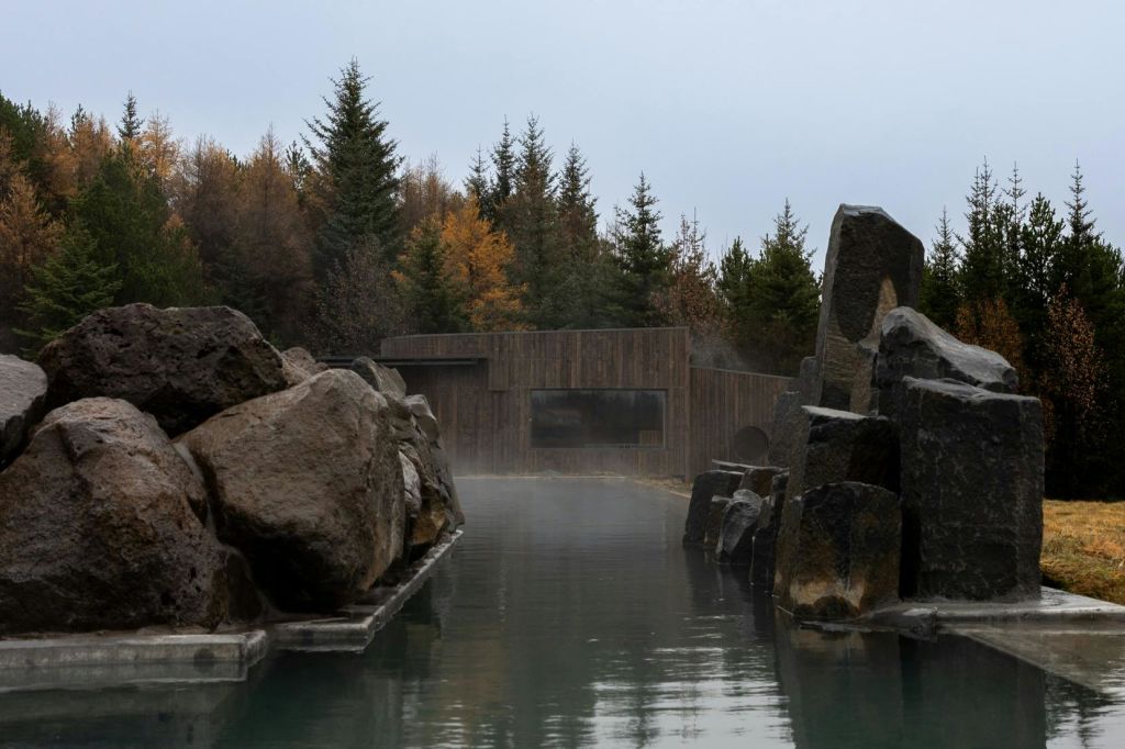 커다란 바위와 소나무로 둘러싸인 고요한 온천을 배경으로 목조 건물이 보입니다.