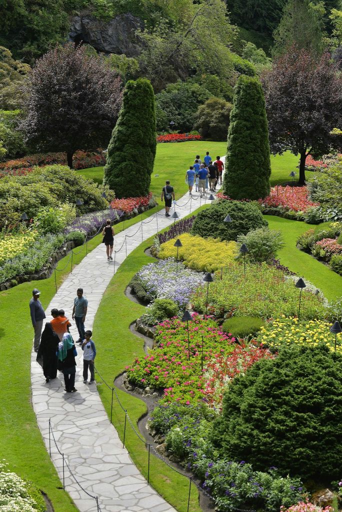 잘 손질된 잔디밭, 화려한 화단, 높은 나무가 있는 활기찬 정원에서 돌길을 따라 산책하는 사람들.