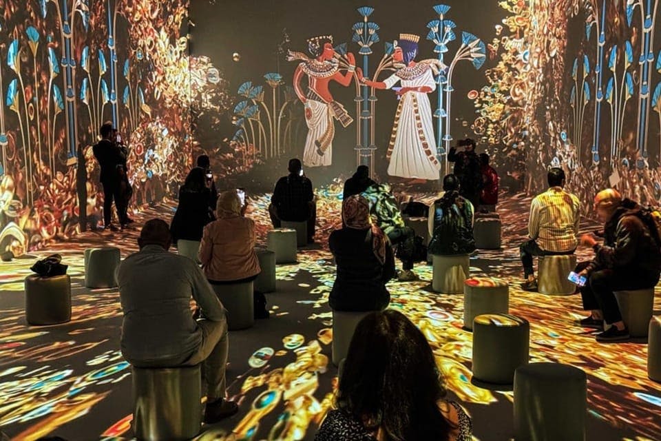 벽과 바닥에 다채로운 고대 이집트 예술 작품이 전시된 조명이 희미한 방에 사람들이 의자에 앉아 있습니다.