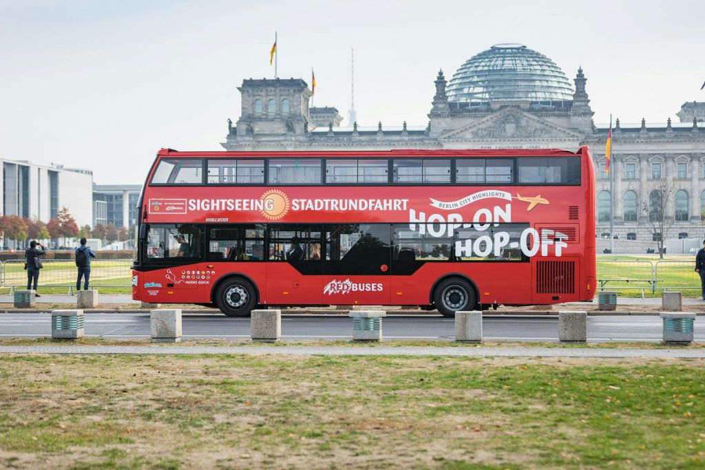 유리 돔이 있는 유서 깊은 건물 앞에 "Hop On Hop Off" 표지판이 있는 빨간색 2층 투어 버스가 있습니다.