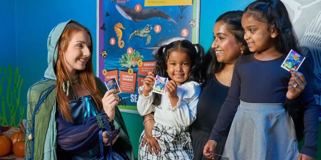 세 명의 어린이와 한 여성이 해양 생물에 대한 다채로운 교육 포스터 옆에서 미소를 짓고 있습니다. 한 아이가 카드를 들고 있습니다.