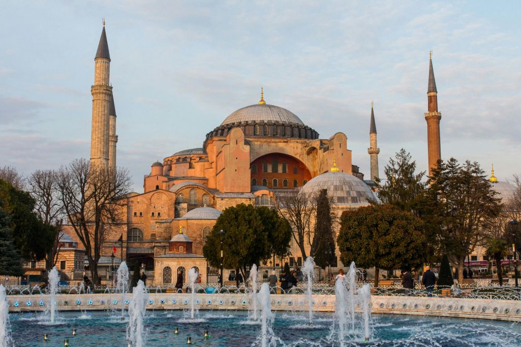 미나레트, 전경의 나무, 부분적으로 흐린 하늘 아래 광장의 분수가 있는 유서 깊은 돔형 건물입니다.