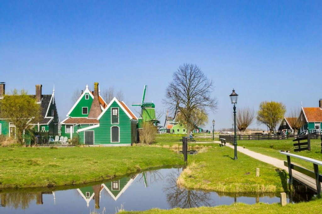 작은 운하 근처의 초록색 주택과 풍차, 통로를 따라 나무와 가로등이 있으며, 모두 맑고 푸른 하늘 아래 있습니다.