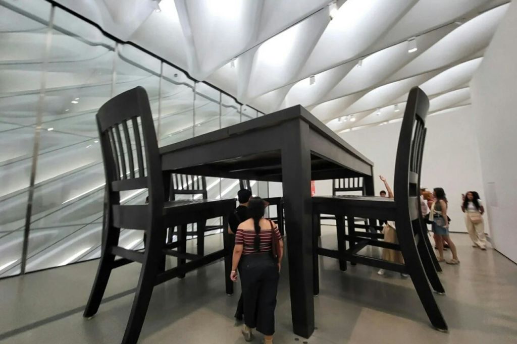 높고 하얀 천장이 있는 현대적인 전시 공간에서 대형 테이블과 의자 아래를 걷는 사람들.