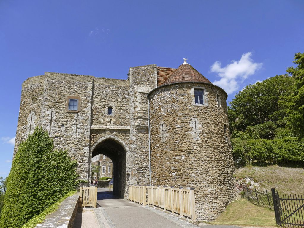 맑고 푸른 하늘 아래 둥근 포탑과 나무로 만든 다리, 주변 녹지가 있는 돌로 만든 성 입구.