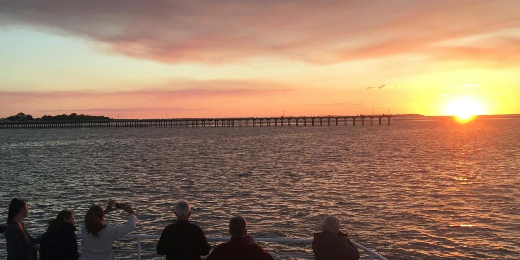 긴 부두를 배경으로 석양이 질 무렵 바다가 내려다보이는 난간 옆에 세 사람이 서 있습니다.