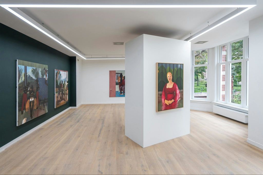 나무 바닥과 흰색 벽이 있는 현대 미술관으로, 중앙 기둥에 빨간 드레스를 입은 여성의 그림이 전시되어 있습니다.