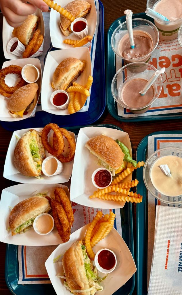 테이블 위에 햄버거, 감자튀김, 양파 링, 디핑 소스, 밀크쉐이크 등 4개의 패스트푸드 트레이가 놓여 있습니다.