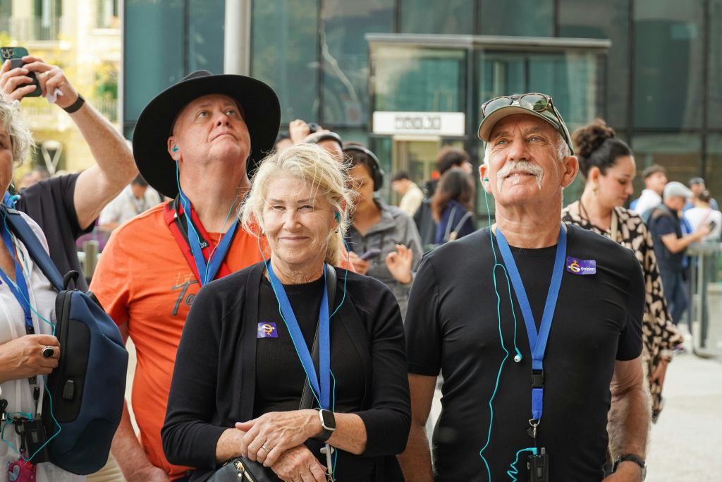 랜야드와 이어폰을 착용한 한 무리의 사람들이 야외에서 현대적인 유리 건물 앞에 서 있습니다.