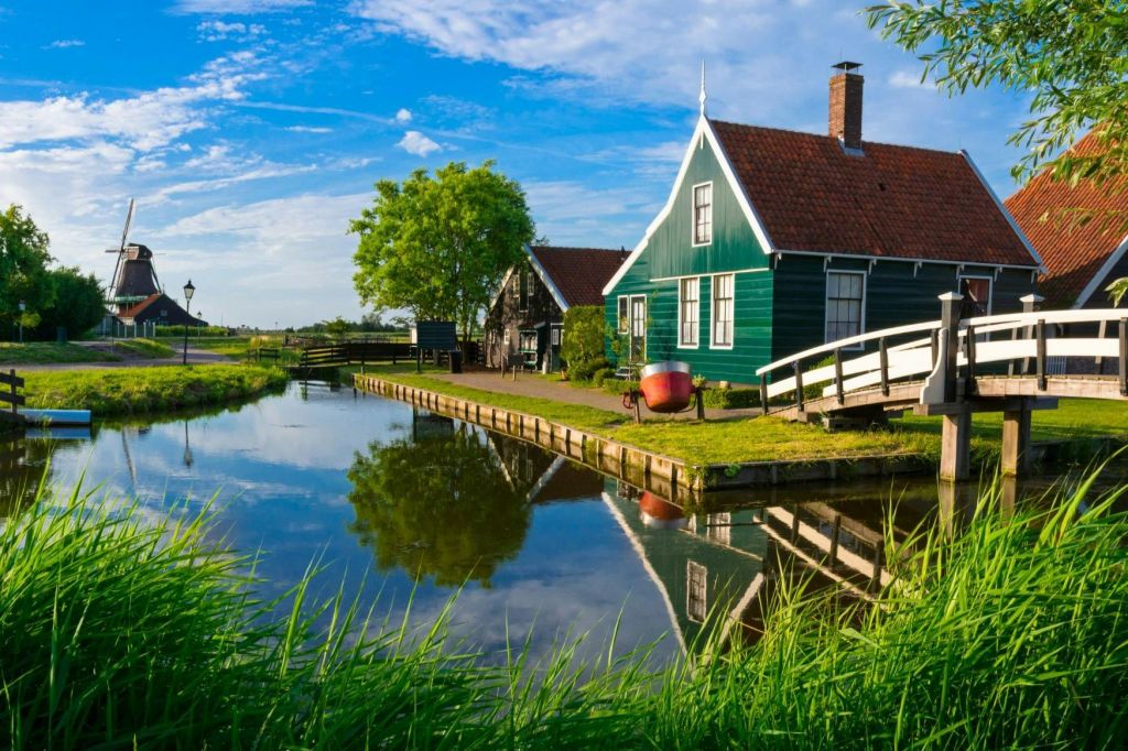 잔잔한 운하 옆의 붉은 지붕, 하얀 풋브릿지, 푸른 하늘 아래 흩어져 있는 구름이 우거진 녹음이 우거진 녹색 주택.