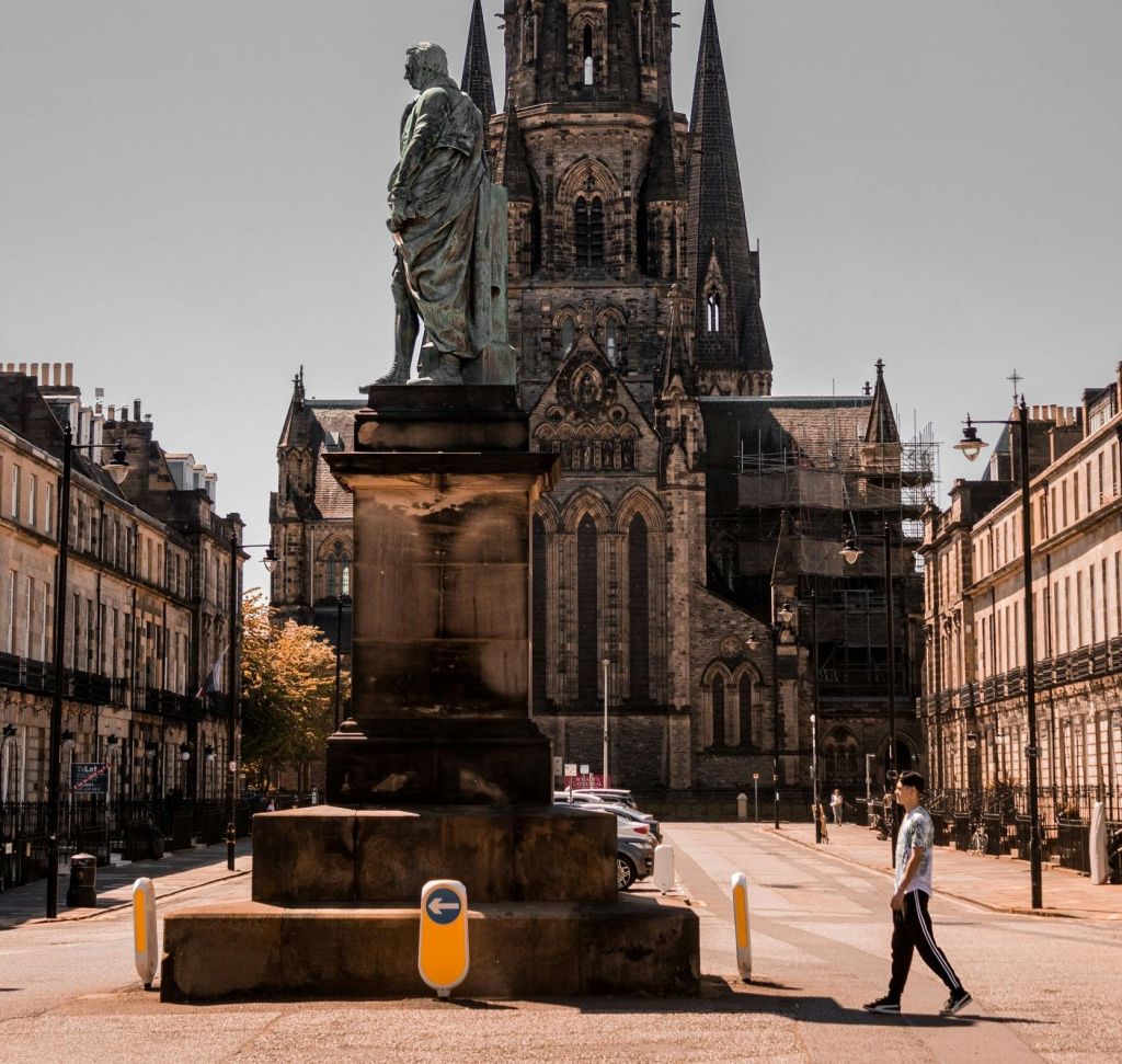 한 사람이 커다란 고딕 양식의 교회를 배경으로 한 도시 광장의 동상 옆을 걸어가고 있습니다.