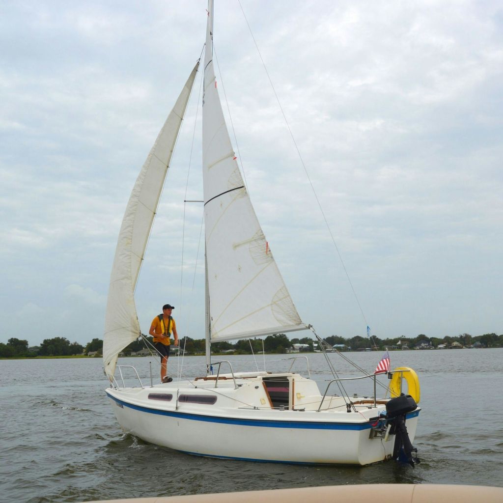 흐린 하늘과 육지를 배경으로 차분한 물 위에 구명조끼를 입은 두 사람이 탄 작은 범선입니다.