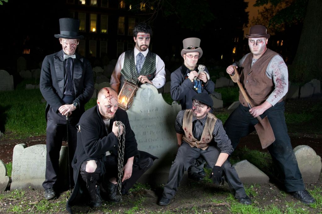 빅토리아 시대 의상을 입은 5명이 묘지에서 포즈를 취하고 있으며, 중앙에 비석이 있는 쇠사슬과 등불과 같은 소품을 들고 있습니다.