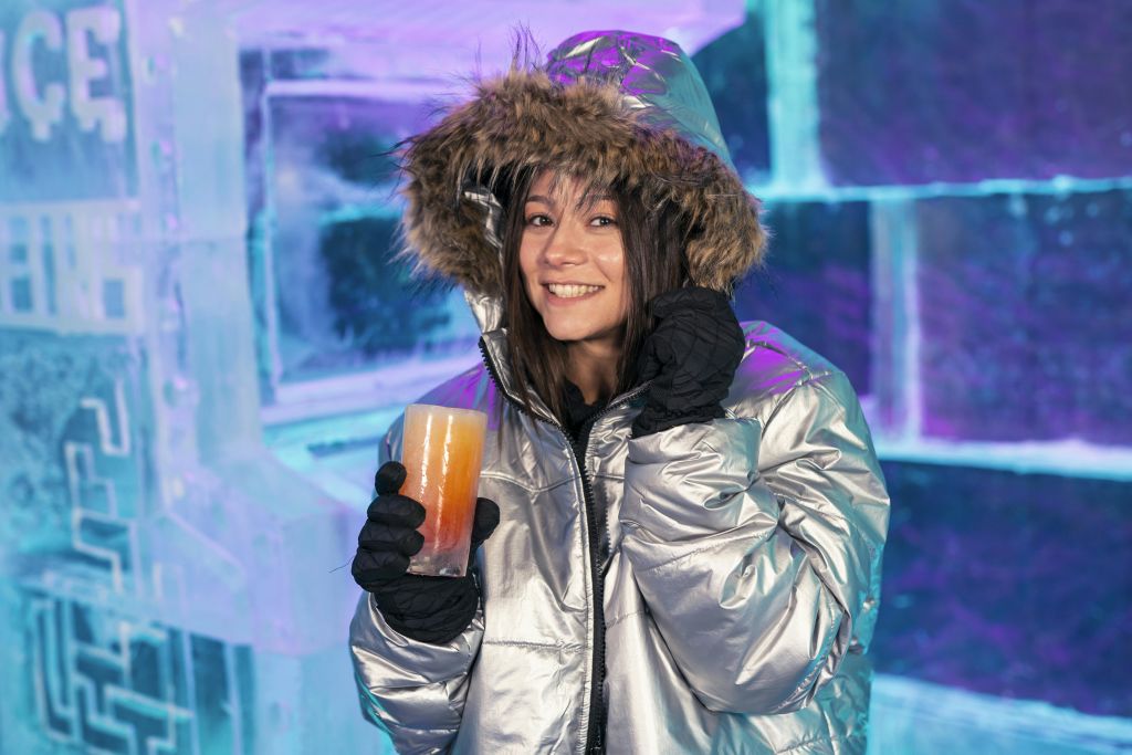 모피를 두른 후드가 달린 은색 파카를 입은 여성이 얼음 유리잔에 음료를 담고 있습니다.