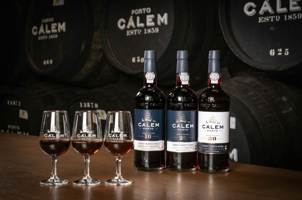 칼렘 포르투 와인 3병과 테이블 위에 놓인 와인잔, 통을 배경으로 한 와인.
