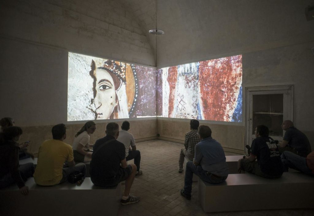 조명이 희미한 방의 벤치에 앉아 두 개의 벽에 놓인 고대 프레스코 스타일 작품의 커다란 투영을 감상하는 사람들.