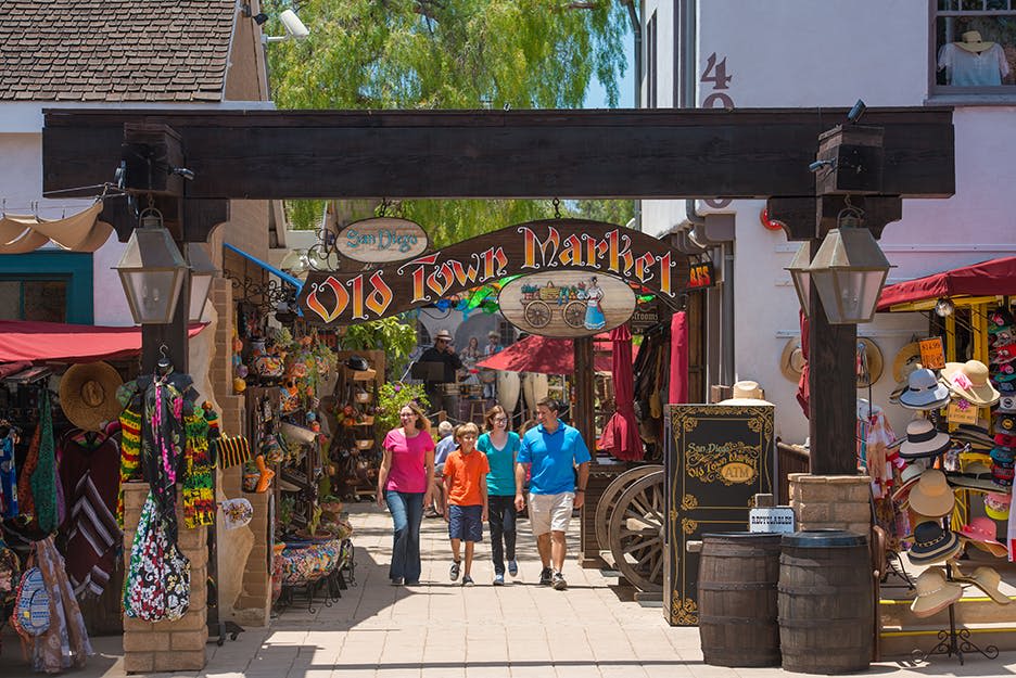 상점, 다채로운 물품, 마차, 통이 있는 시장 구역에서 "올드 타운 마켓" 표지판 아래를 걷는 사람들의 그룹.